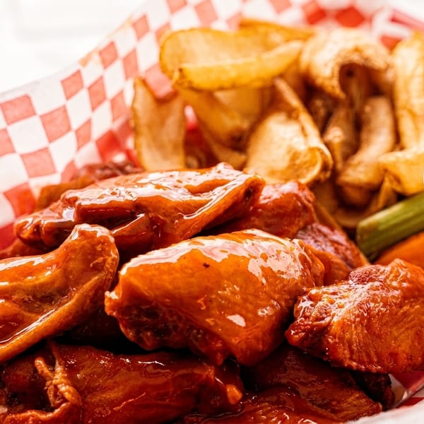 A red and white checkered basket filled with glazed chicken wings, potato wedges, and ranch dressing.