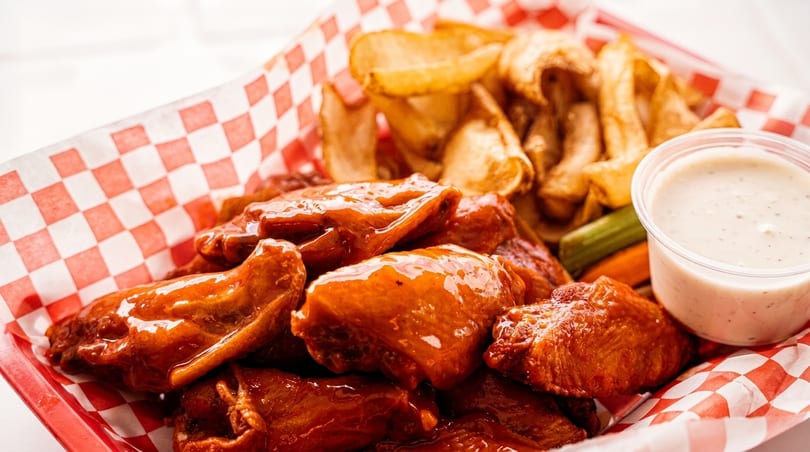 A red and white checkered basket filled with glazed chicken wings, potato wedges, and ranch dressing.