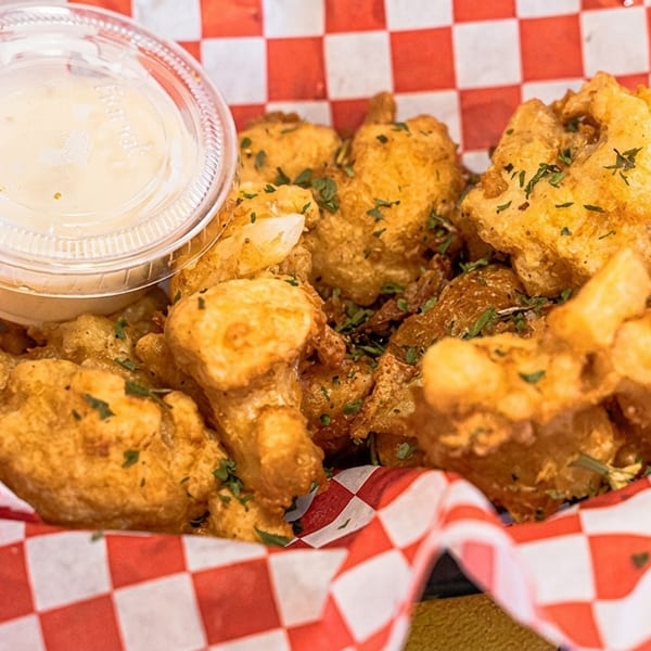 Golden fried cheese curds with parsley flakes and a side of dipping sauce in a red and white checkered basket.