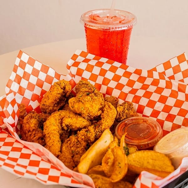 Fried chicken and potato wedges in a basket with red checkered paper, alongside a red drink.