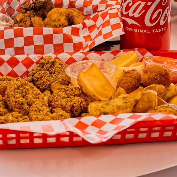 Two red baskets filled with fried chicken, potato wedges, and dipping sauces on a white table.